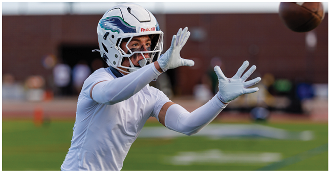 A person wearing a white football uniform and gloves is reaching out with both hands as if to catch a ball on a sports field. Description generated by AI