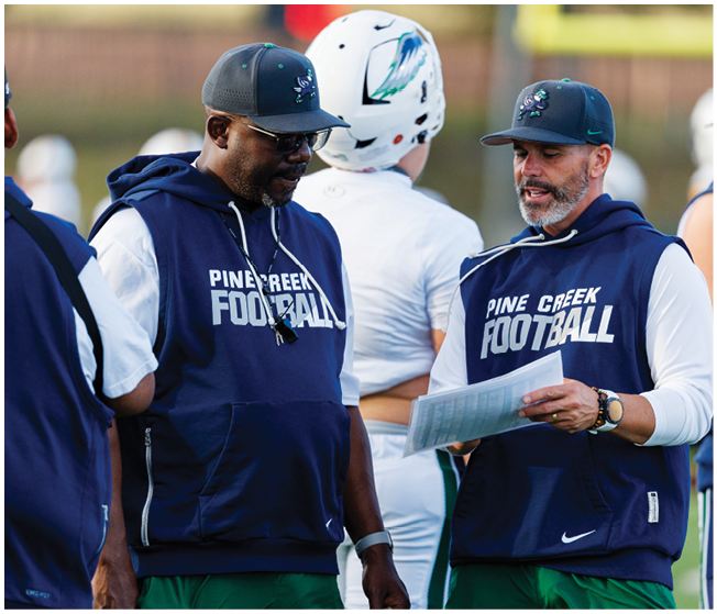 Two people wearing Pine Creek Football hoodies and caps are standing outdoors, with one holding a paper and the other looking at it. Description generated by AI