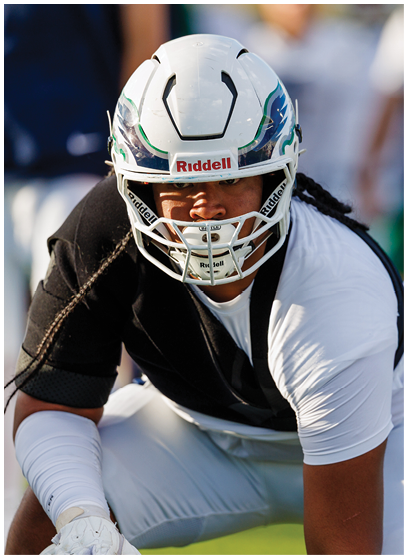 A person wearing a white football helmet and black and white athletic clothing is crouched in a ready position on a field. Description generated by AI