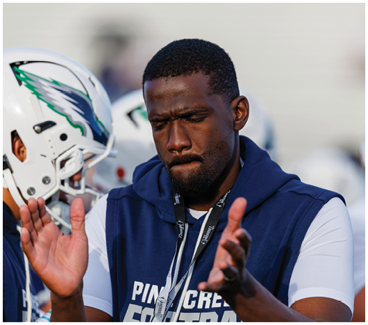 A person wearing a navy blue hoodie with a whistle around their neck is clapping while standing next to another person in a football helmet. Description generated by AI