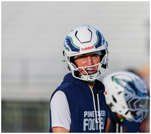 A person wearing a football helmet and a sleeveless hoodie with “Pine Creek Football" printed on it is standing outdoors with another person in a football helmet in the background. Description generated by AI