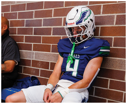 A person in a football uniform and helmet sits against a brick wall looking down with hands clasped. Description generated by AI