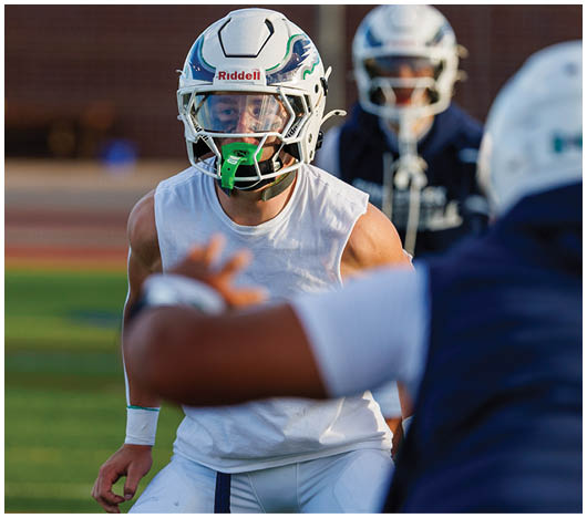 A person wearing a white sleeveless shirt and football helmet with a green mouthguard is focused and ready on a sports field while two other people in helmets and sports gear are in the background. Description generated by AI
