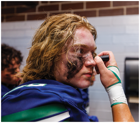 A person with long hair wearing a blue sports jersey is applying black face paint under their eyes in a locker room. Description generated by AI