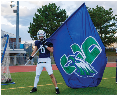 A person in a football uniform and helmet is holding a large blue flag with a green and white eagle logo on a sports field while another person stands nearby. Description generated by AI