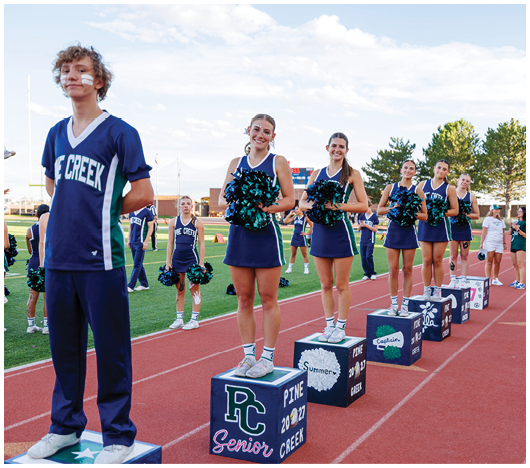 A group of people in cheerleading uniforms stand on decorated blocks on a running track with pom-poms, smiling and posing during an outdoor event. Description generated by AI