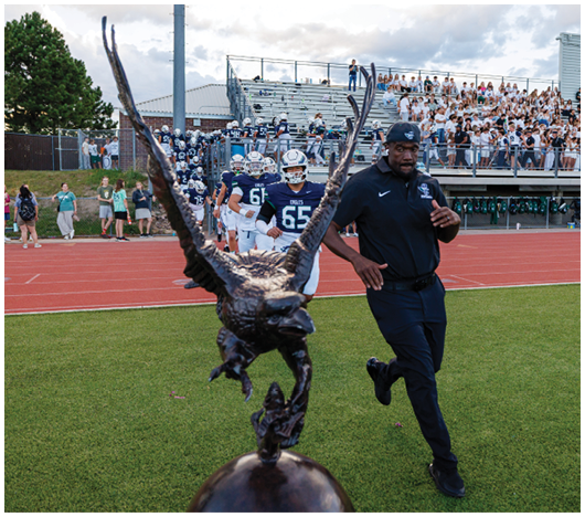 A person in a dark outfit runs on a sports field near a large eagle statue with a group of football players and spectators in the background. Description generated by AI