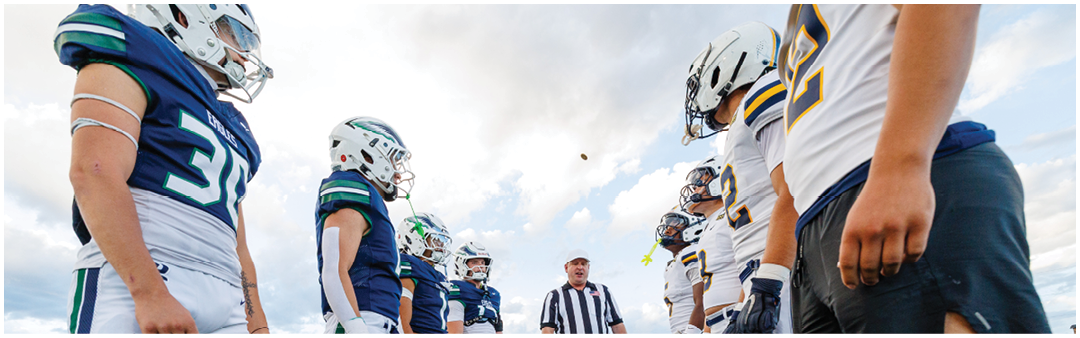 Two opposing football teams in uniforms stand facing each other on a field while a referee in a striped shirt prepares to toss the ball. Description generated by AI
