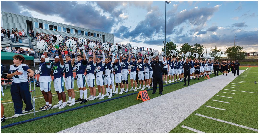 A line of people in matching sports uniforms stand on a football field holding helmets raised in the air with a crowd in the bleachers and a cloudy sky in the background. Description generated by AI