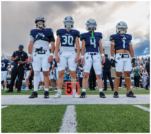 Four football players in uniforms stand on a field next to a marker with the number five while people and other players are visible in the background. Description generated by AI