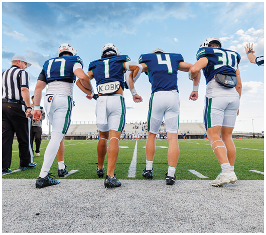 Four football players in blue and white uniforms stand arm in arm on a field with two referees nearby under a partly cloudy sky. Description generated by AI