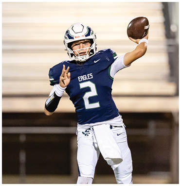 A person wearing a blue football uniform with the number two is preparing to throw a football on a field with blurred bleachers in the background. Description generated by AI