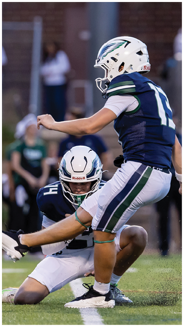 Two people in football uniforms are on a field with one holding the ball in place while the other kicks it. Description generated by AI
