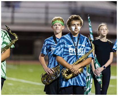 Two people wearing blue tie-dye shirts and holding saxophones stand on a field, with one person wearing a green headband and the other having white face paint. Description generated by AI
