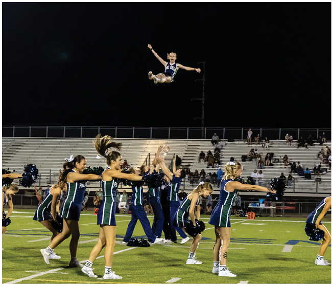 A group of cheerleaders in uniform perform a routine on a football field at night with one cheerleader being thrown high into the air above the others. Description generated by AI
