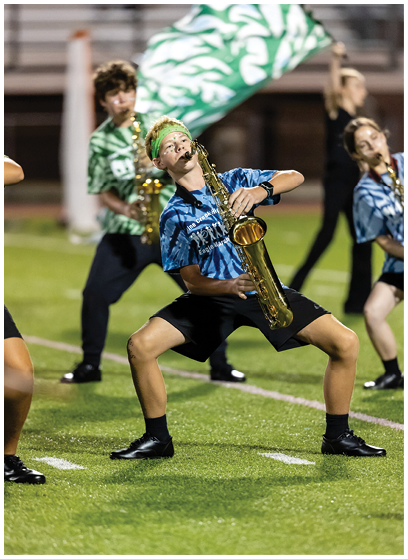 A person wearing a blue tie-dye shirt and green headband is playing a saxophone outdoors with a green and white flag in the background. Description generated by AI