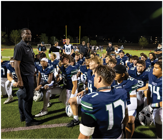 A group of people in football uniforms are gathered on a field at night listening attentively to a person standing and speaking to them. Description generated by AI