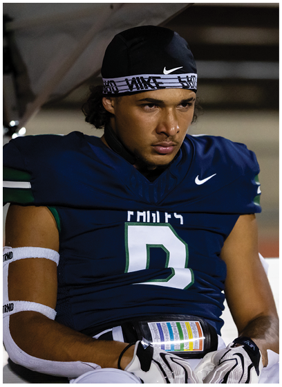 A person wearing a dark blue football uniform with the number 9 and a black Nike headband is sitting on a bench looking focused. Description generated by AI
