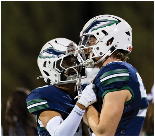 Two people wearing football helmets and blue jerseys are facing each other closely, with one person holding the other's jersey in an outdoor setting. Description generated by AI