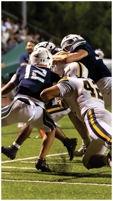 Three people in football uniforms are engaged in a tackle on a grassy field during a game. Description generated by AI