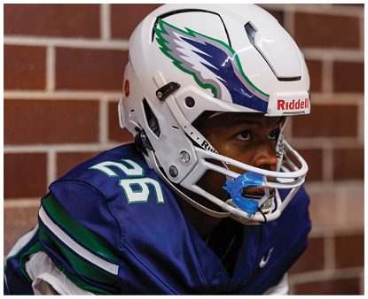 A person wearing a blue and green football uniform and white helmet is sitting against a brick wall, looking focused. Description generated by AI