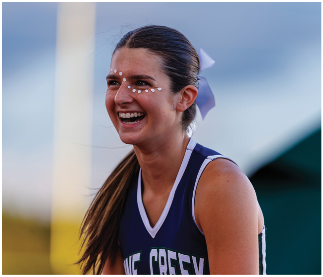 A person wearing a sports uniform with a ponytail and face paint is smiling outdoors with a blurred background. Description generated by AI