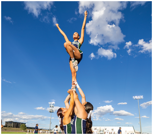 A person in a cheerleading uniform is being lifted into the air by others against a bright blue sky with scattered clouds. Description generated by AI