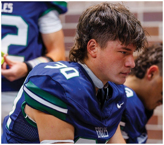 A person wearing a blue football jersey with the number 30 sits on a bench looking focused, with another person in a similar jersey partially visible in the background. Description generated by AI