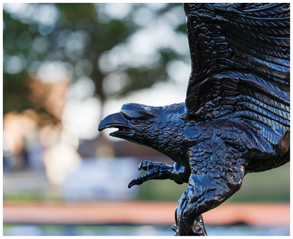 A close-up of a detailed dark metal eagle sculpture with wings spread and talons extended against a blurred outdoor background. Description generated by AI