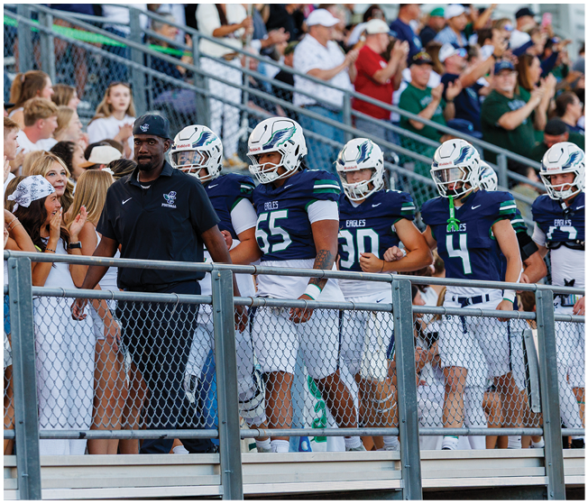 A group of people in football uniforms and helmets stand on bleachers next to a person in a black polo shirt and cap while a crowd watches in the background. Description generated by AI