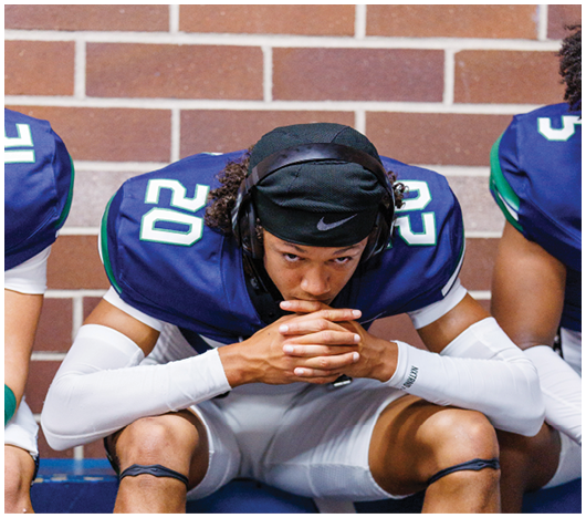 A person wearing a blue and white football uniform with the number 20 is sitting on a bench with hands clasped and head slightly bowed against a brick wall background. Description generated by AI