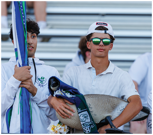 Two people stand in front of bleachers, one holding a flag and the other wearing sunglasses and a backwards cap while holding a large decorative goose wrapped in a scarf. Description generated by AI
