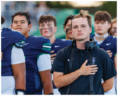 A group of people in sports uniforms stand with one person in front wearing a headset and placing a hand over their chest. Description generated by AI