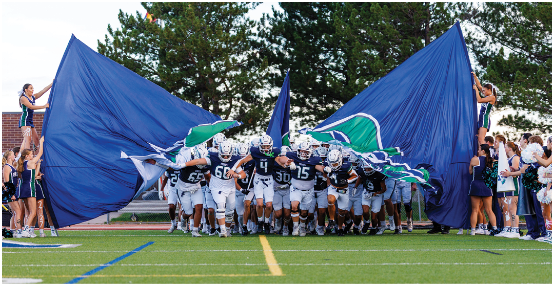 A group of football players in white uniforms run onto the field through a large blue banner held open by people on either side. Description generated by AI