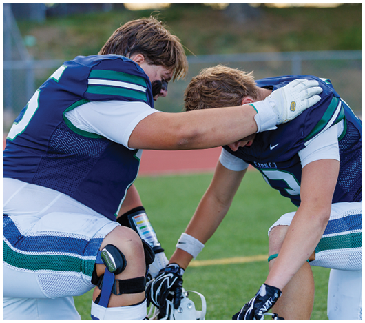 Two people in football uniforms kneel on a field with one person comforting the other by placing a hand on their shoulder. Description generated by AI
