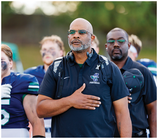 A group of people in football uniforms and a person wearing a headset and a polo shirt with a football logo stand together outdoors, with some placing their hands over their hearts. Description generated by AI