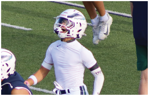 Two people in white sports uniforms stand on a football field, with one person clapping and the other facing away. Description generated by AI