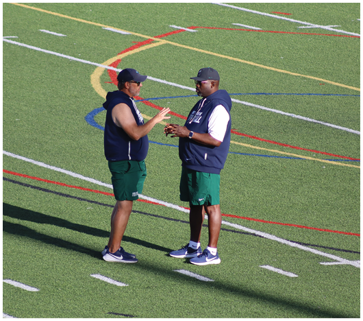 Two people wearing white football helmets with red faceguards and a red lion logo are focused and looking ahead. Description generated by AI