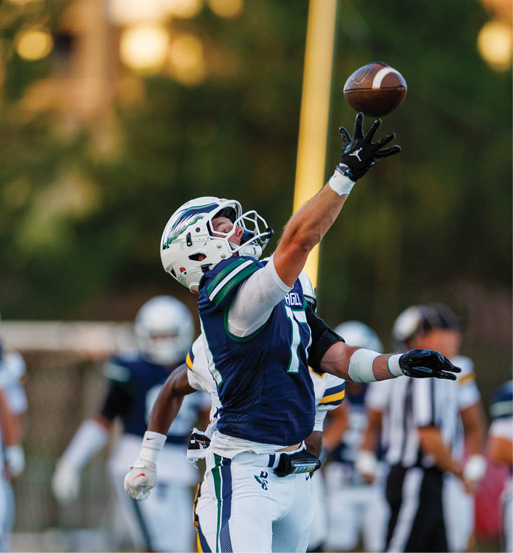 A person in a football uniform and helmet is reaching up with one hand as if to catch or block a ball during a game, with other players and a referee visible in the background. Description generated by AI