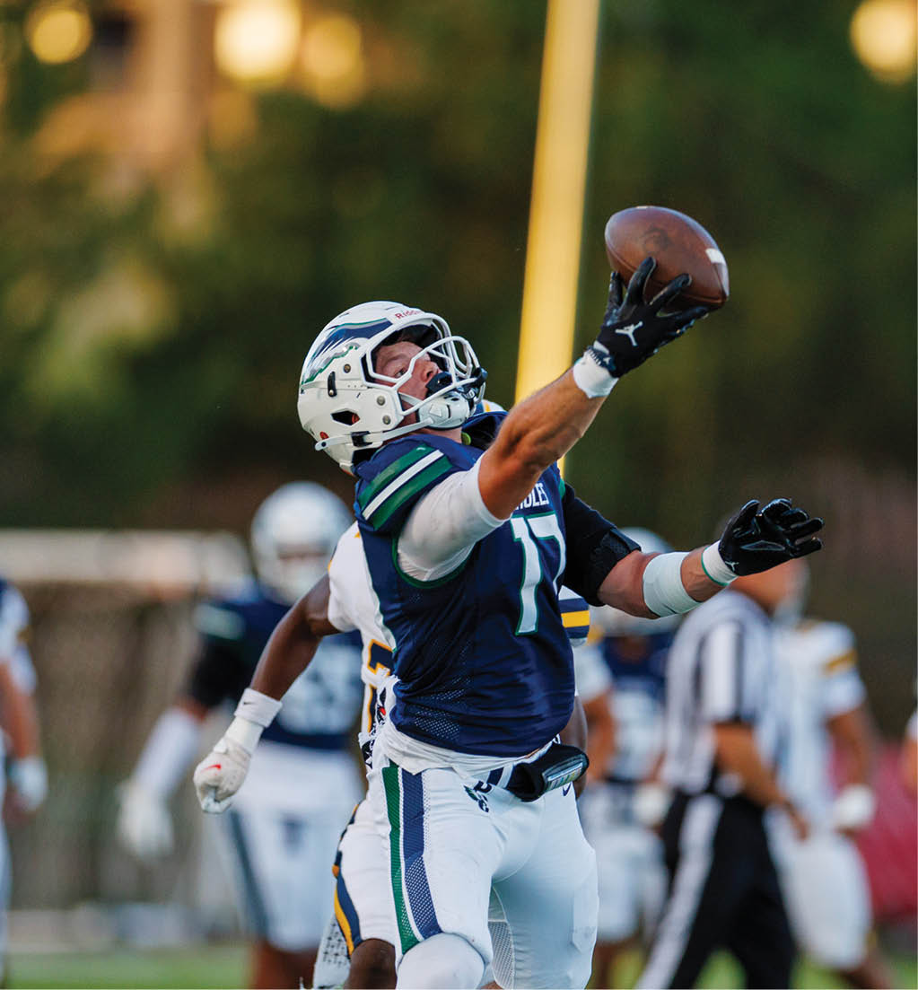 A person in a football uniform is reaching up with one hand to catch a football during a game with other players and a referee visible in the background. Description generated by AI