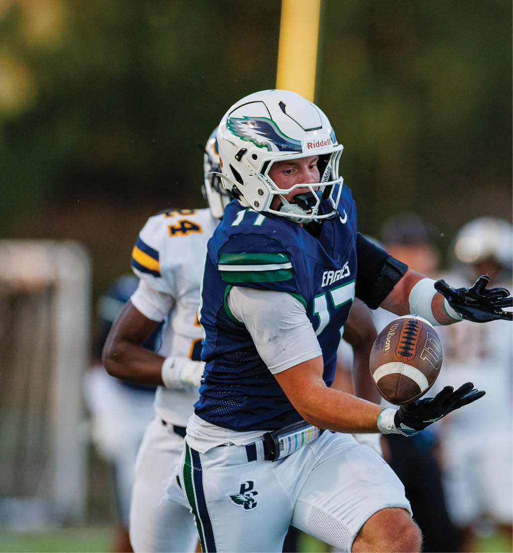 A person wearing a blue and white football uniform with a helmet is catching a football while another person in a white and yellow uniform is behind them on a field. Description generated by AI