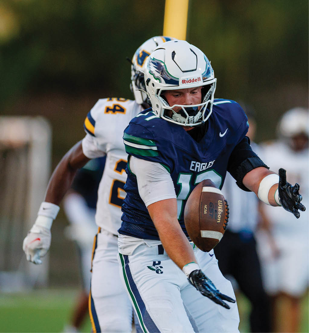 A person in a blue and white football uniform with “Eagles" on the jersey is catching a football while another person in a white and yellow uniform is behind them on a grassy field. Description generated by AI