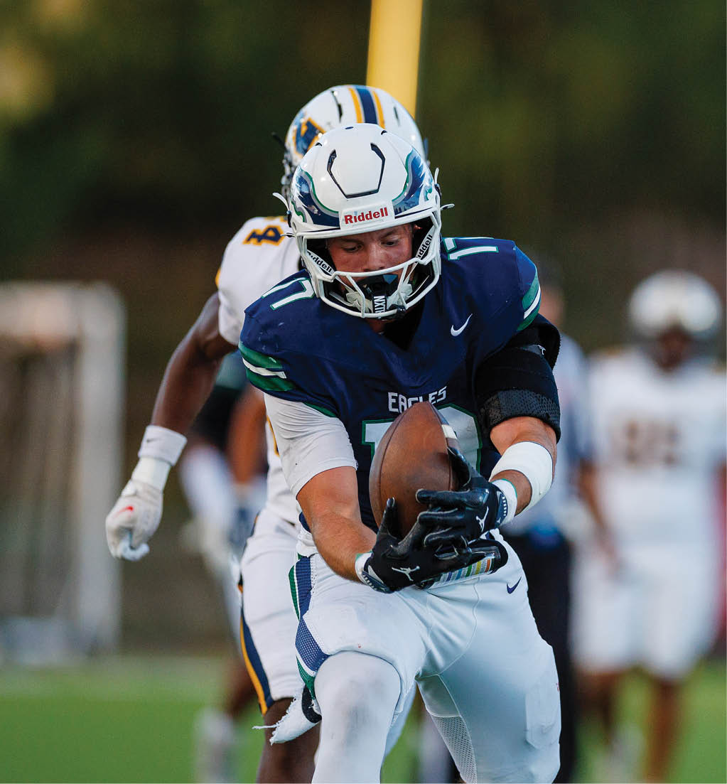 A person in a blue and white football uniform is catching a football while another person in a white and yellow uniform is closely behind on a grassy field. Description generated by AI