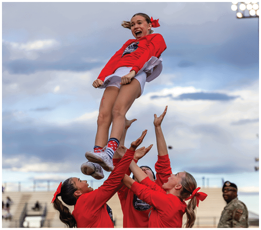 A group of people in red cheerleading uniforms are performing a stunt outdoors with one person being lifted high into the air against a cloudy sky. Description generated by AI