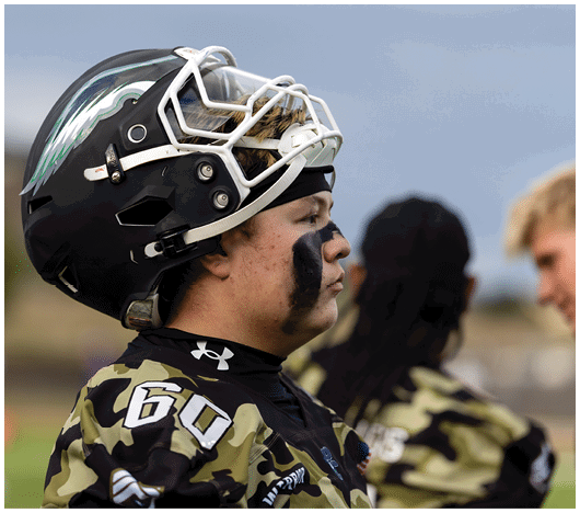 A person wearing a football helmet and camouflage jersey with face paint stands in profile while two other people in similar jerseys are blurred in the background. Description generated by AI