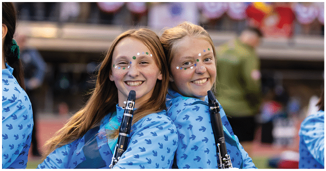 Two smiling people with face paint and blue patterned shirts hold clarinets while standing back to back at an outdoor event. Description generated by AI