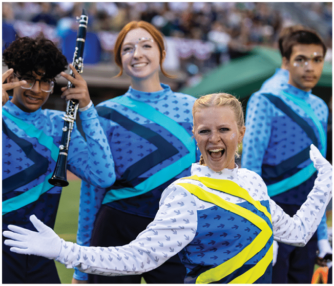 A group of people in colorful marching band uniforms are smiling and posing, with one person holding a clarinet and another enthusiastically extending their arms. Description generated by AI