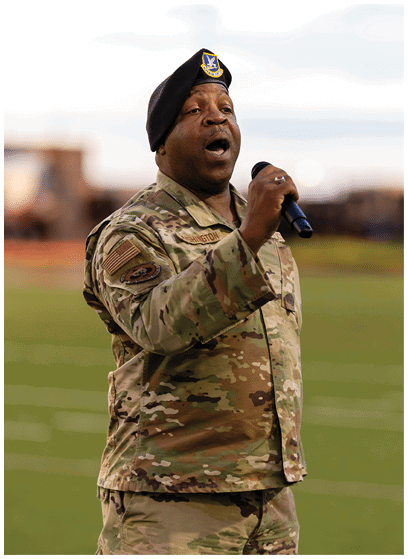 A person in military uniform and beret is holding a microphone and appears to be singing or speaking on a field. Description generated by AI
