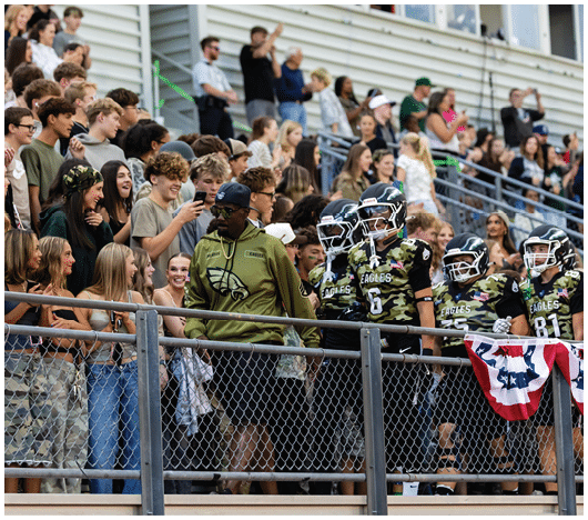 A large group of people wearing casual and camouflage clothing are gathered closely together in bleachers, some smiling and interacting with each other. Description generated by AI