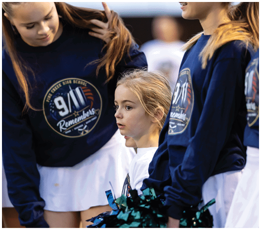 Children wearing matching 9/11 remembrance sweatshirts and white skirts stand together, with one child in the center holding green and black pom-poms. Description generated by AI